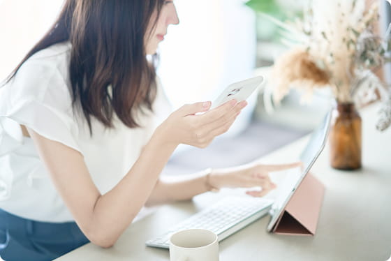 白いブラウスの女性がスマートフォンを持ちながらタブレットを操作している